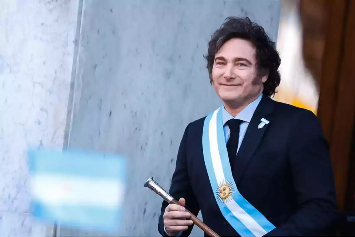 Un hombre con traje oscuro y corbata negra lleva una banda presidencial celeste y blanca con un sol dorado, mientras sostiene un bastón de mando frente a un fondo de mármol.