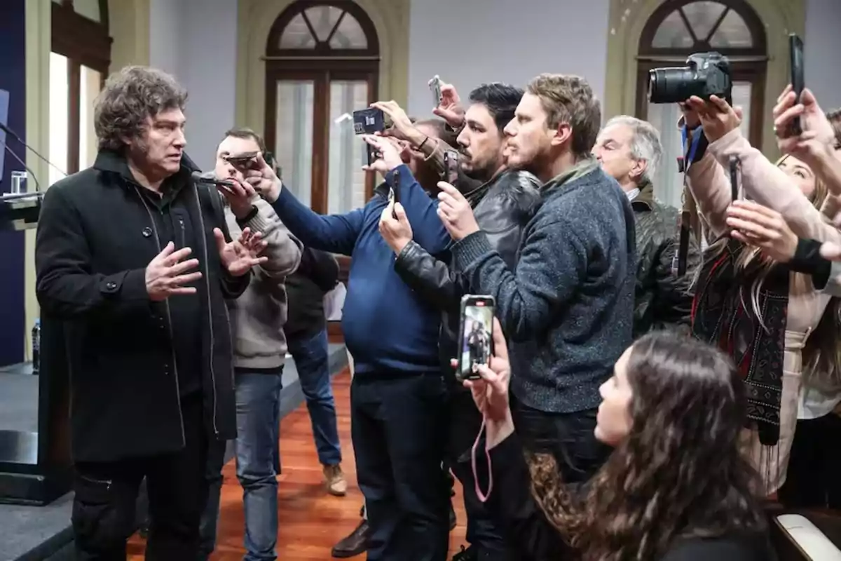 A man is speaking while a group of people surrounds him, holding cameras and mobile phones to record or take pictures. A man is speaking while a group of people surrounds him, holding cameras and mobile phones to record or take pictures.
