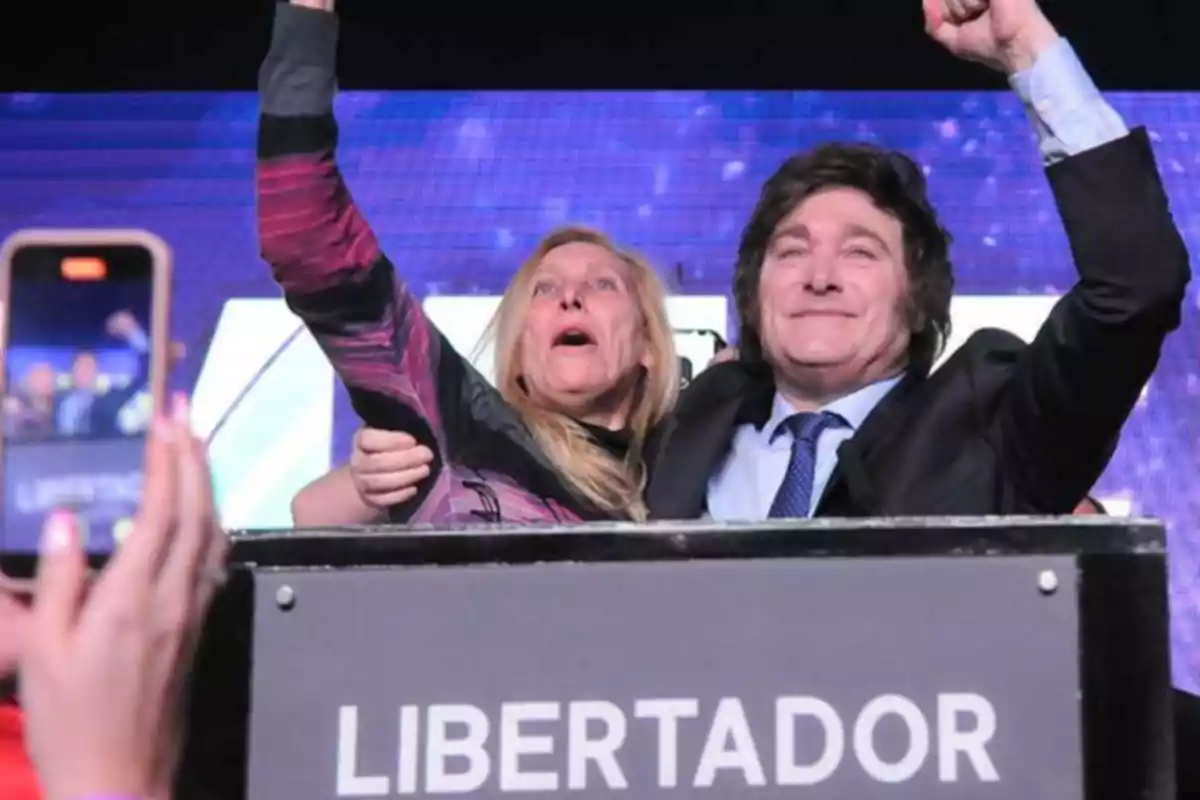 Dos personas celebrando en un escenario con un cartel que dice "LIBERTADOR" y una multitud tomando fotos.