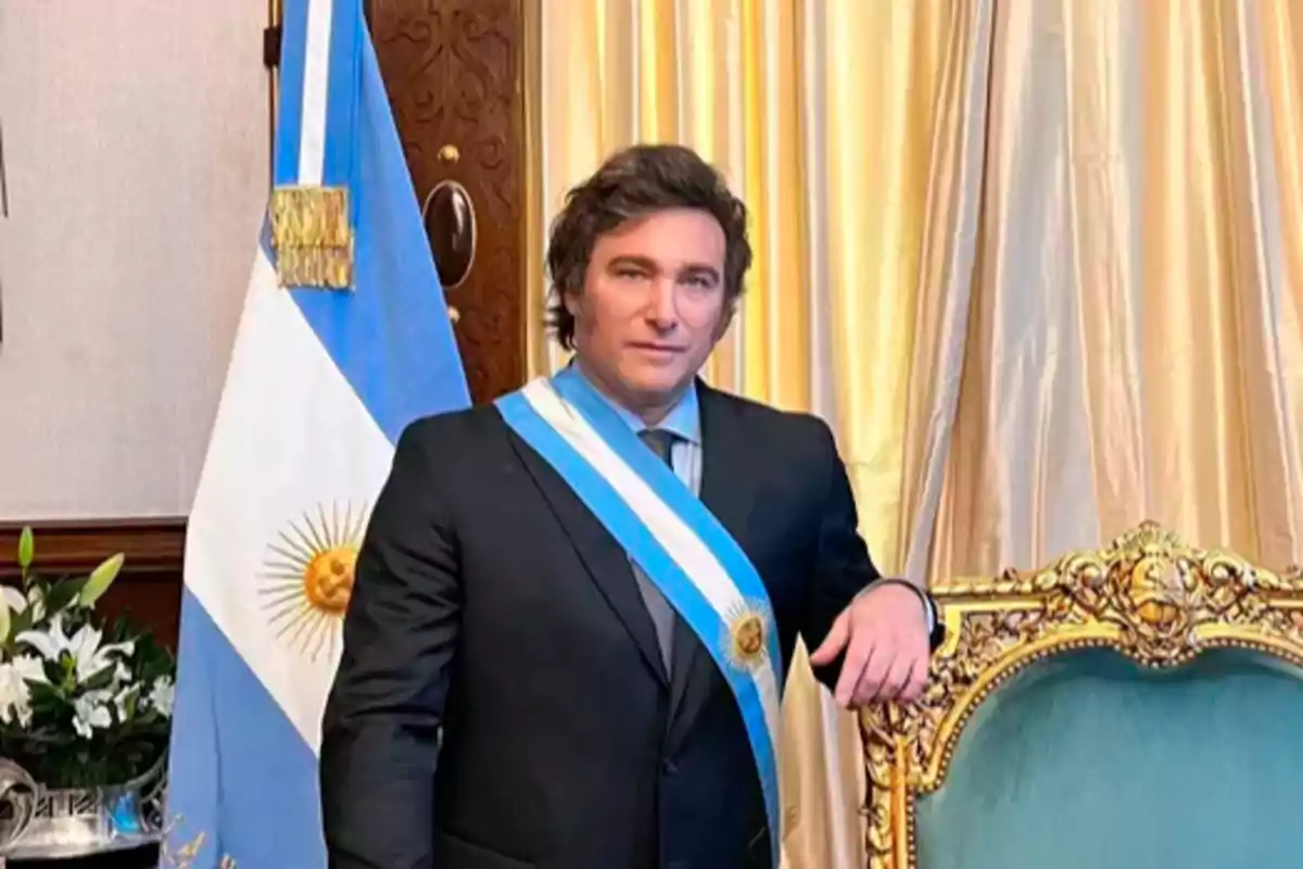 Un hombre con traje y banda presidencial posa junto a una silla decorativa y una bandera argentina en un entorno elegante.