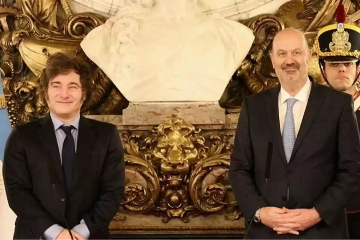 Dos hombres de pie en un entorno decorativo con un busto de mármol y un guardia con uniforme ceremonial.