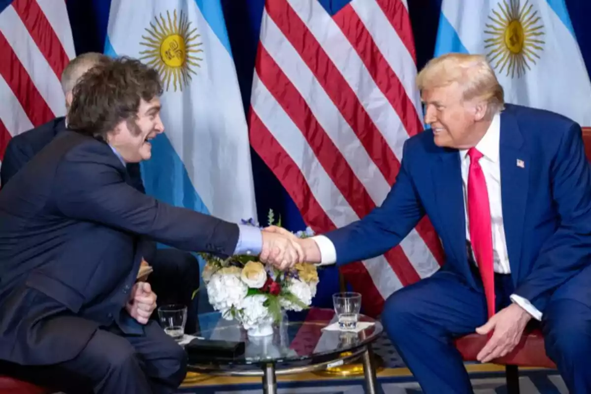 Dos personas dándose la mano en una reunión, con banderas de Argentina y Estados Unidos de fondo.