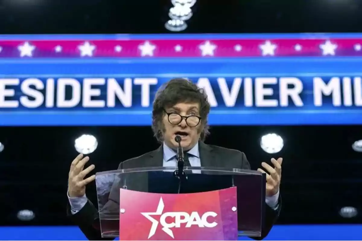 Un hombre hablando en un podio durante un evento de CPAC con un cartel azul de fondo.