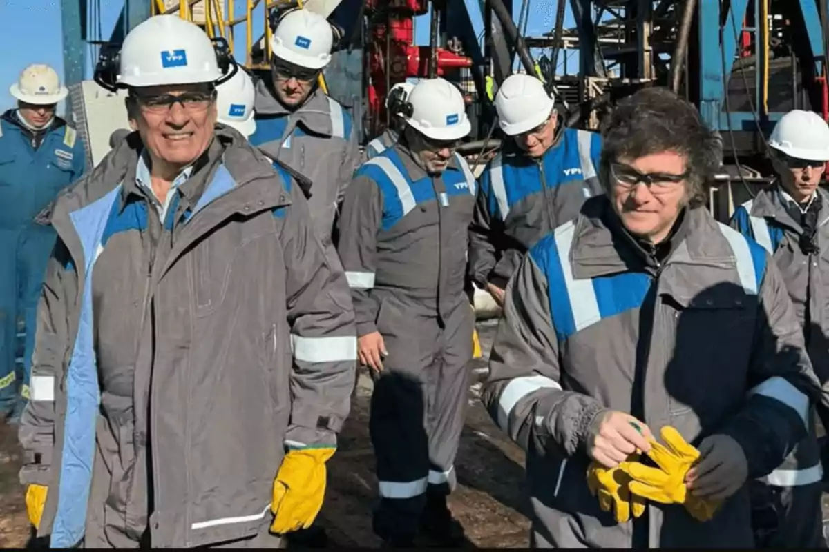 Un grupo de personas con cascos y ropa de trabajo industrial, algunos con guantes amarillos, se encuentran en un entorno de maquinaria pesada.