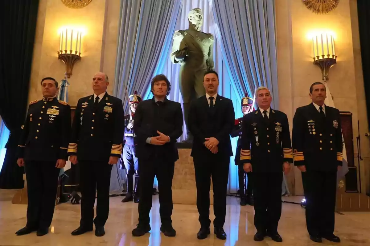 Un grupo de hombres en uniforme militar y traje formal posan frente a una estatua en un salón decorado con cortinas azules y candelabros.