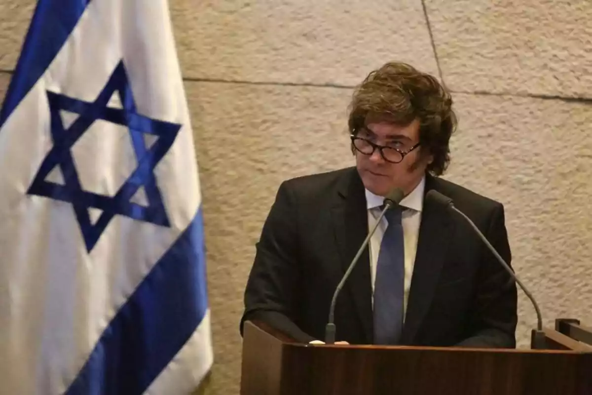 Man in a suit speaking at a podium with an Israeli flag in the background Man in a suit speaking at a podium with an Israeli flag in the background