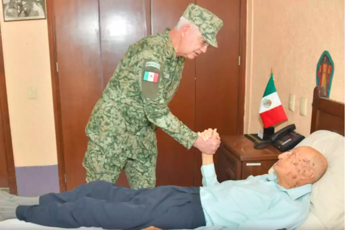 Un militar de uniforme camuflado saluda a un hombre mayor que está acostado en una cama con una bandera de México en el fondo. Un militar de uniforme camuflado saluda a un hombre mayor que está acostado en una cama con una bandera de México en el fondo.