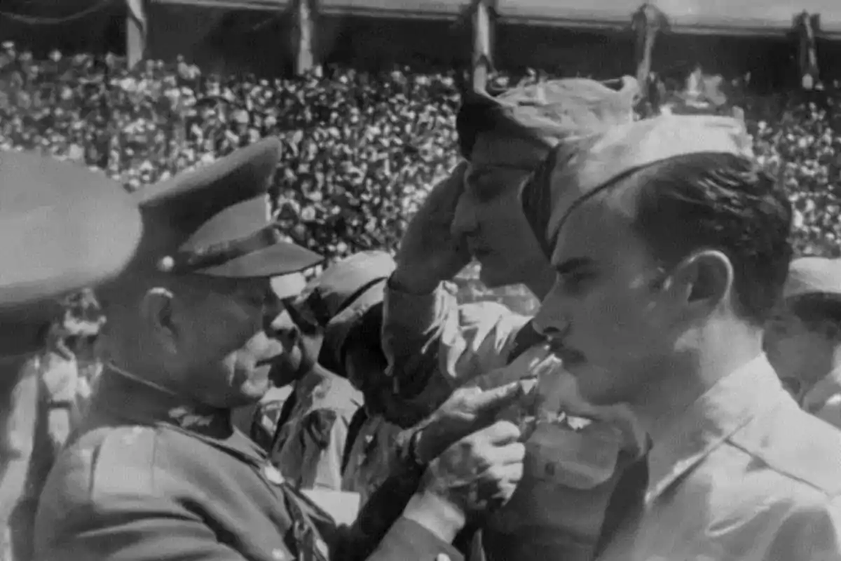 Un oficial militar entrega una medalla a un soldado mientras otro saluda en un evento con una multitud de fondo. Un oficial militar entrega una medalla a un soldado mientras otro saluda en un evento con una multitud de fondo.