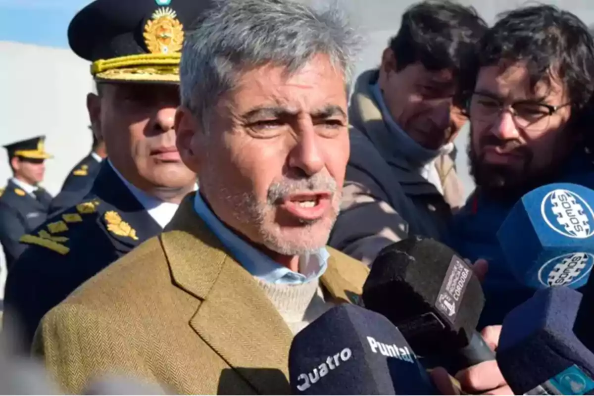 Un hombre hablando frente a varios micrófonos con personas y un oficial de policía en el fondo.