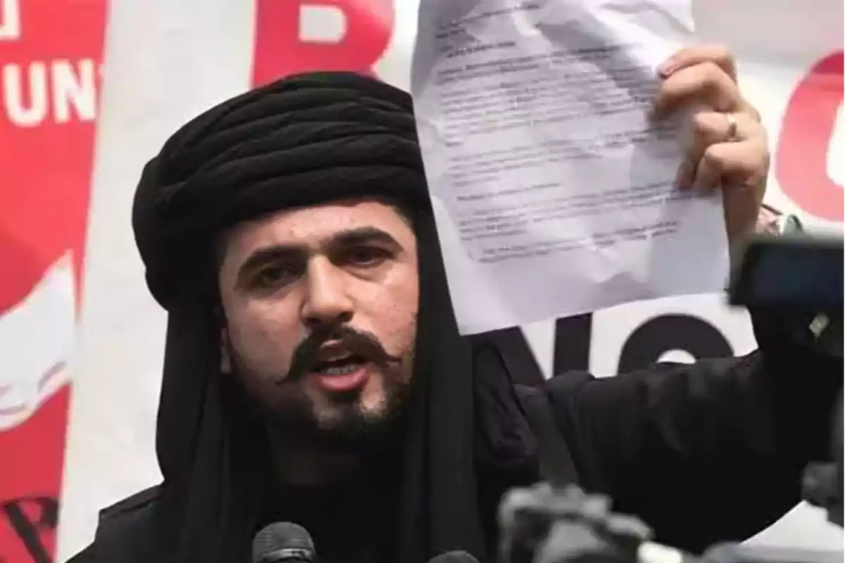 A man with a black turban holds a paper while speaking in front of several microphones. A man with a black turban holds a paper while speaking in front of several microphones.