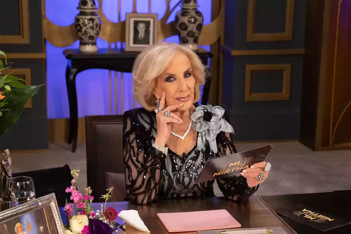 Una persona mayor elegantemente vestida está sentada en una mesa decorada con flores y sostiene una tarjeta mientras mira hacia la cámara.