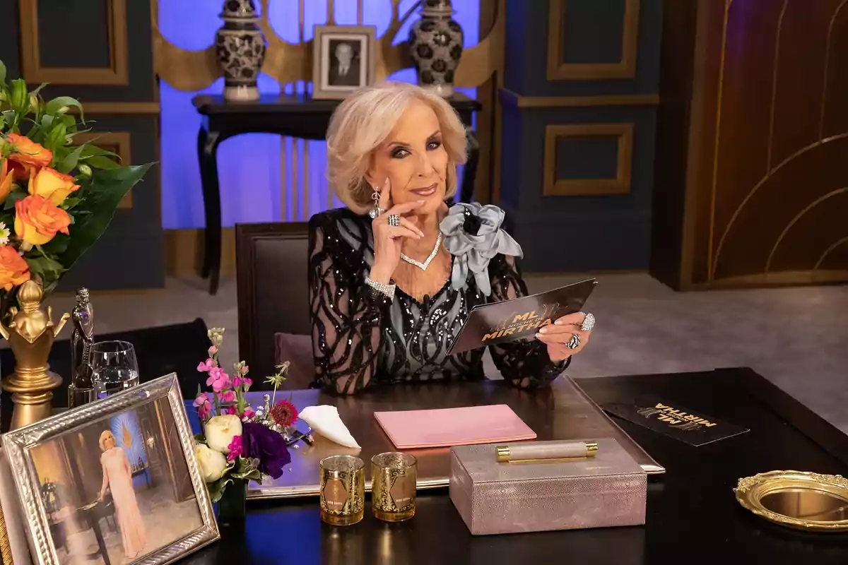 Una persona mayor elegantemente vestida está sentada en una mesa decorada con flores y fotografías, sosteniendo una tarjeta en un set de televisión.