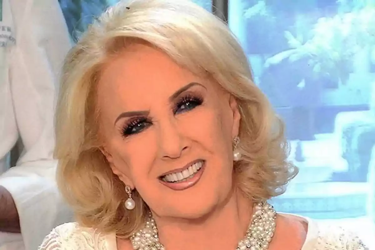 Una persona mayor con cabello rubio y sonrisa, usando joyas y maquillaje elegante, en un entorno iluminado.