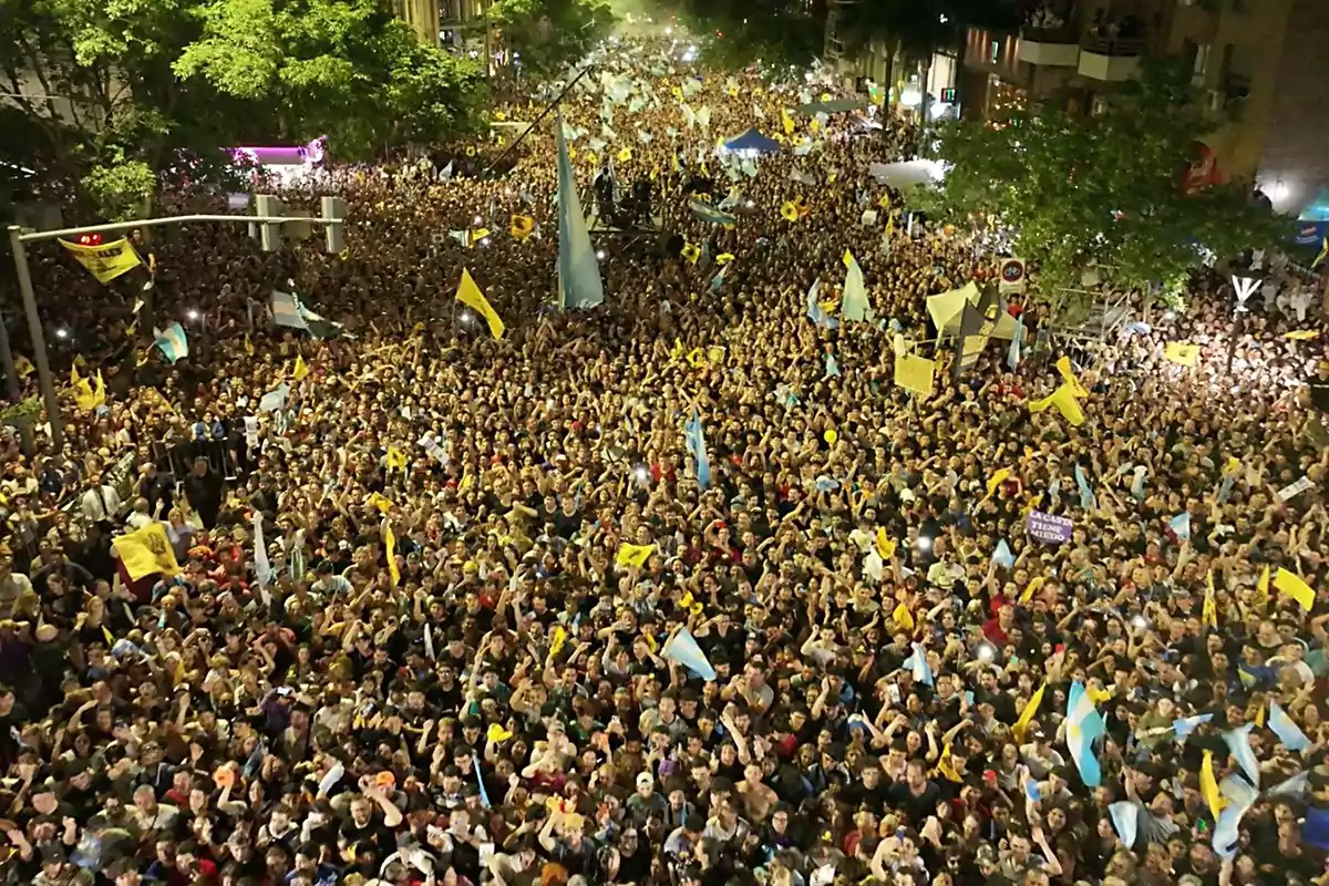 Crowd of people gathered at a nighttime street protest, many of them holding flags and banners, surrounded by trees and city lights. Crowd of people gathered at a nighttime street protest, many of them holding flags and banners, surrounded by trees and city lights.