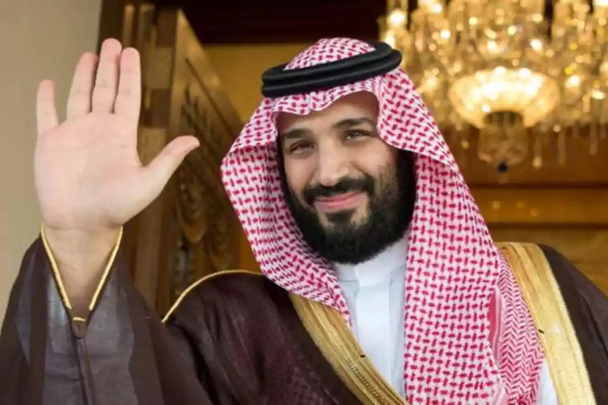 Hombre con barba y vestimenta tradicional árabe saludando con la mano levantada y sonriendo frente a una lámpara de araña Hombre con barba y vestimenta tradicional árabe saludando con la mano levantada y sonriendo frente a una lámpara de araña