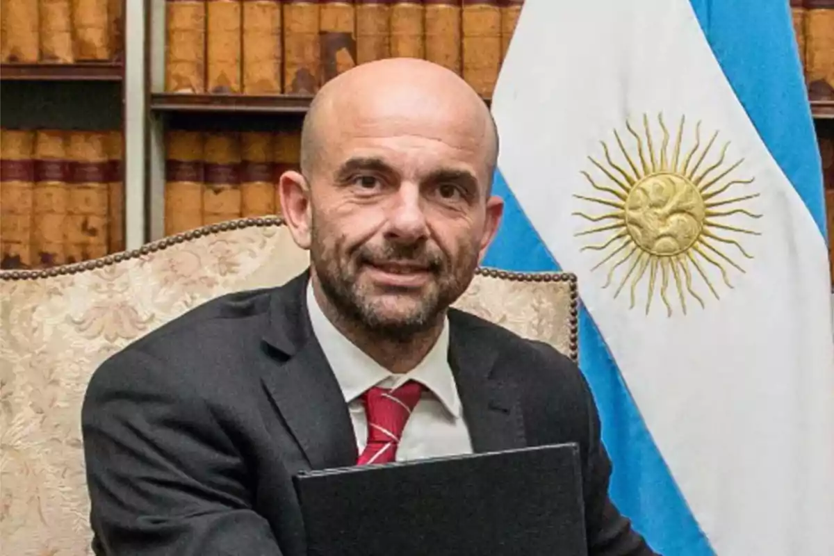 Bald man with a beard and a dark suit sitting in front of an Argentine flag and a bookshelf filled with old books Bald man with a beard and a dark suit sitting in front of an Argentine flag and a bookshelf filled with old books