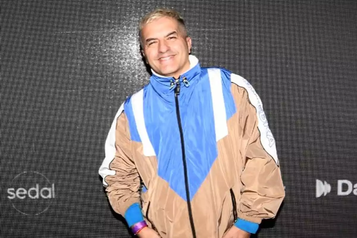 Short-haired blond man smiling, wearing a blue, beige, and white sports jacket, posing in front of a dark background with logos.
