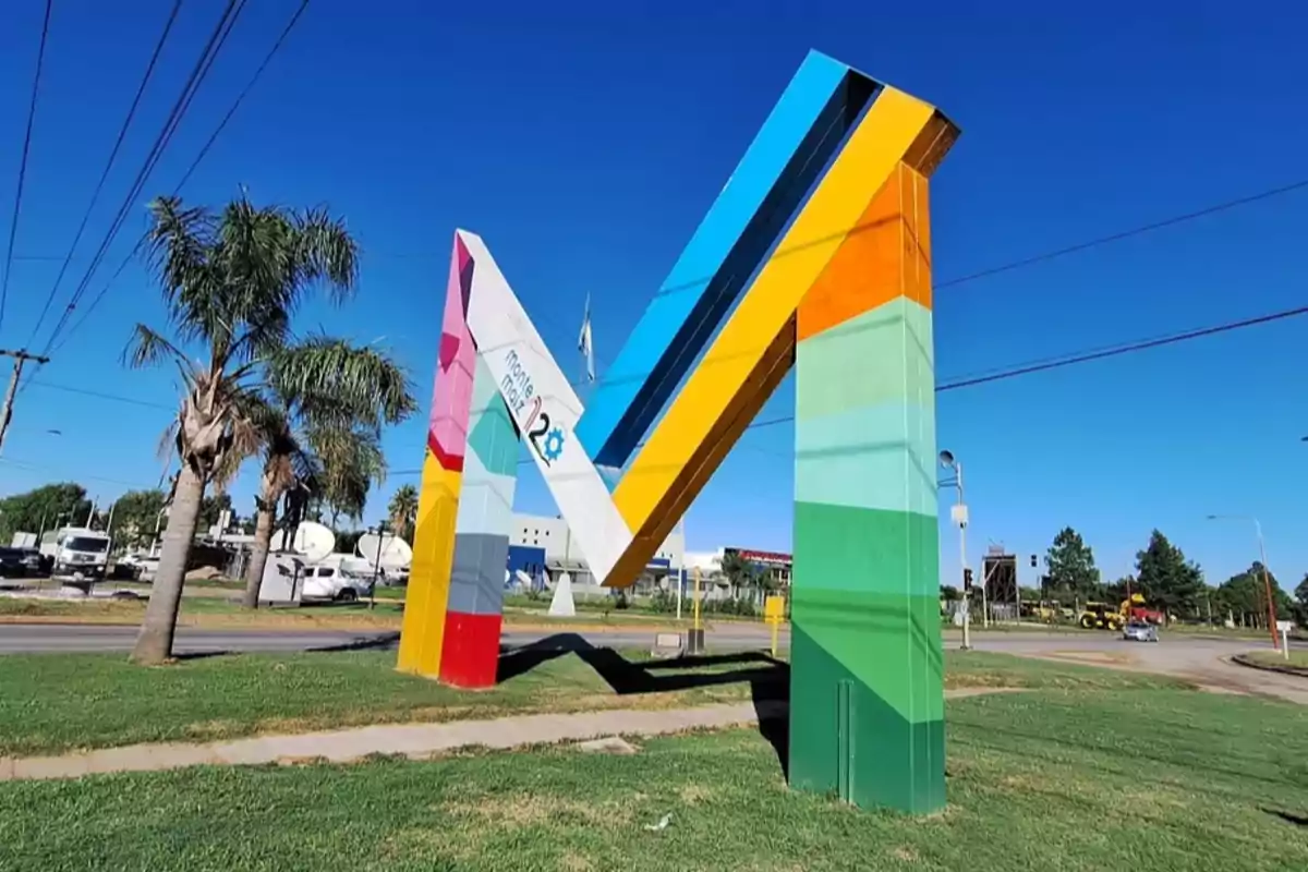 Monumental sculpture in the shape of the letter M painted in bright colors, located on a roundabout with grass and palm trees under a clear sky.