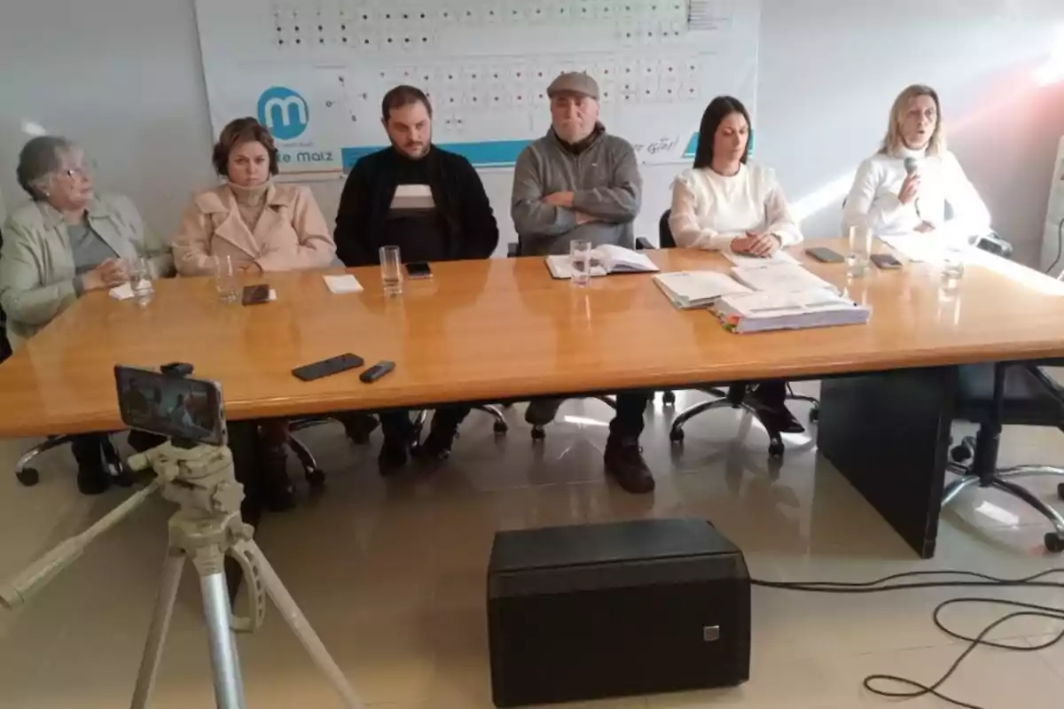 Seis personas sentadas en una mesa de reuniones con documentos, vasos de agua y un micrófono, mientras una cámara en un trípode graba la escena.