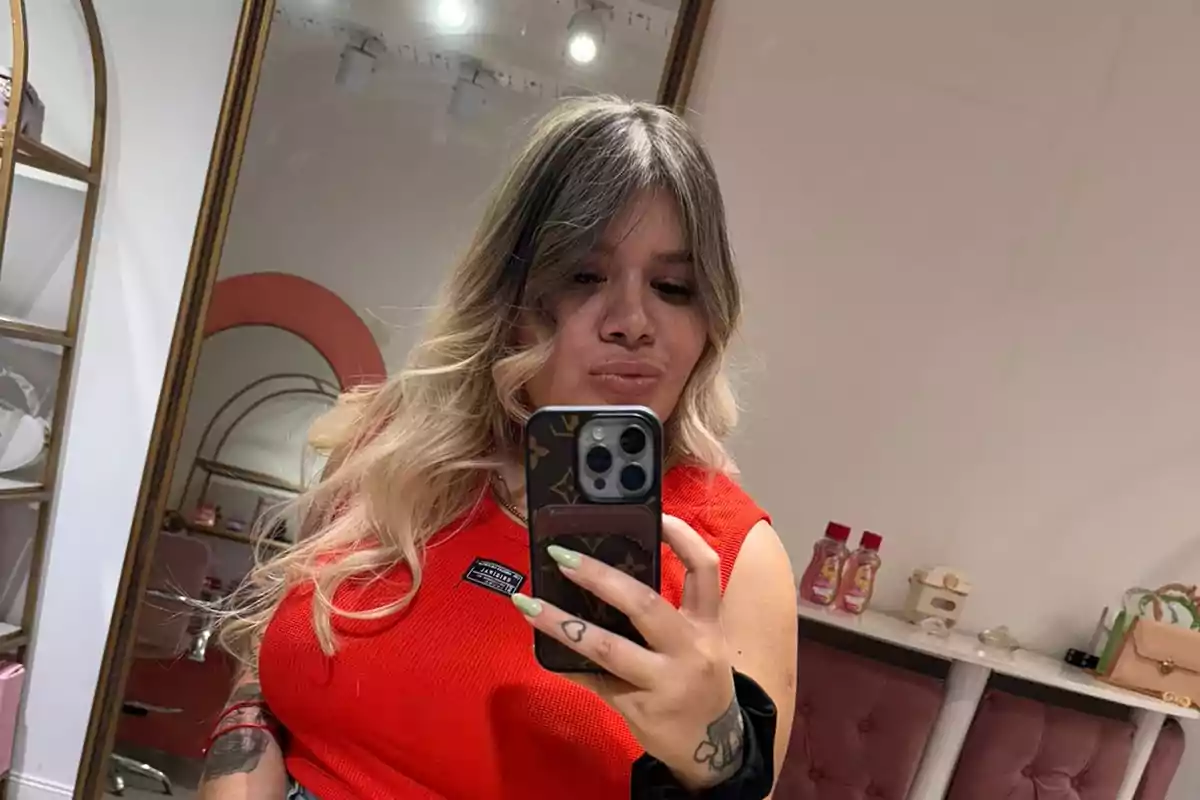 Persona con cabello rubio y oscuro tomándose una selfie frente a un espejo en una habitación decorada con estantes y productos.