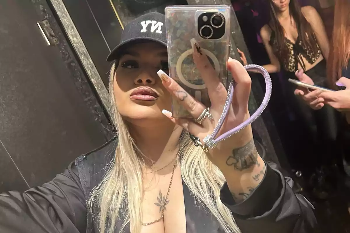 Mujer rubia con gorra negra y chaqueta oscura tomándose una selfie en un baño, con dos mujeres al fondo, una de ellas usando un top de estampado animal y ambas con teléfonos móviles.