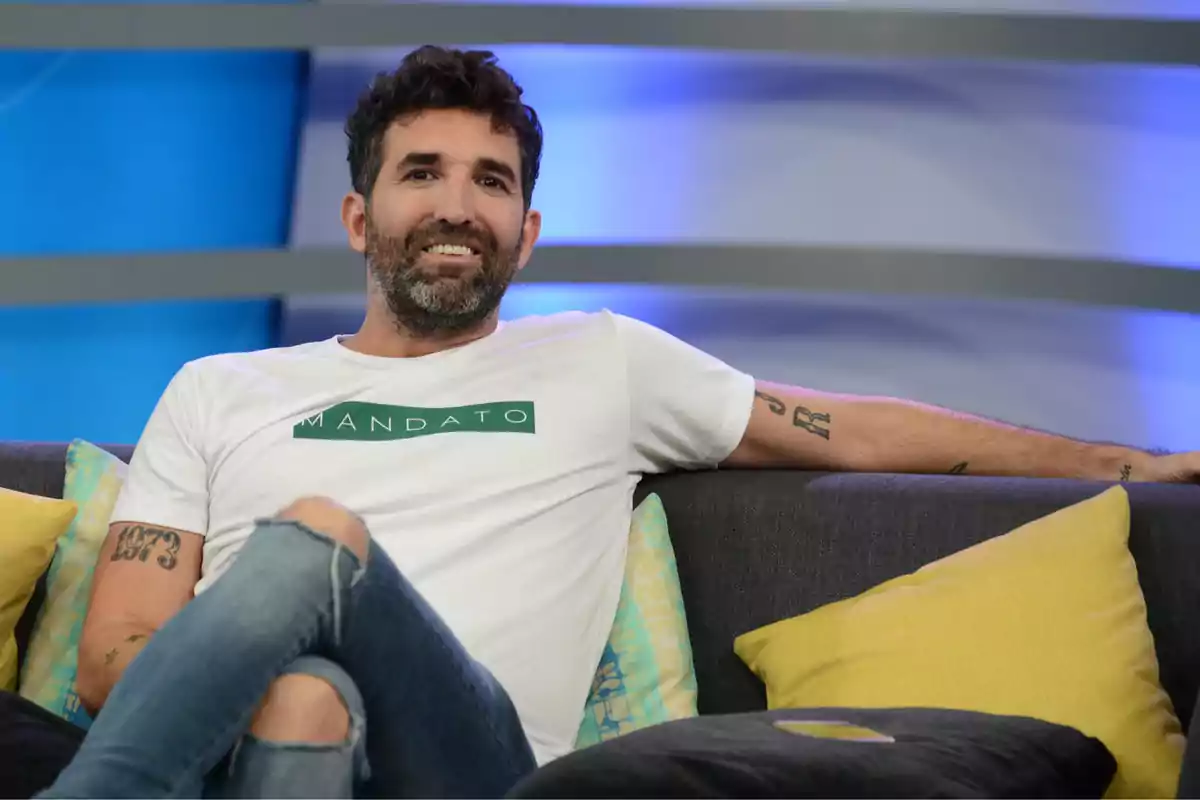 Un hombre con barba y cabello oscuro está sentado en un sofá gris con cojines amarillos y azules, lleva una camiseta blanca con la palabra Un hombre con barba y cabello oscuro está sentado en un sofá gris con cojines amarillos y azules, lleva una camiseta blanca con la palabra