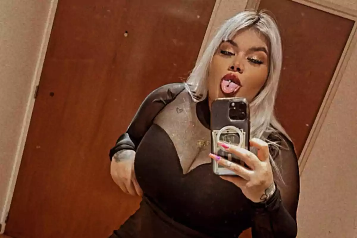 Una persona con cabello rubio y maquillaje llamativo toma una selfie frente a un espejo mostrando la lengua y un piercing, usando un vestido negro con transparencias.