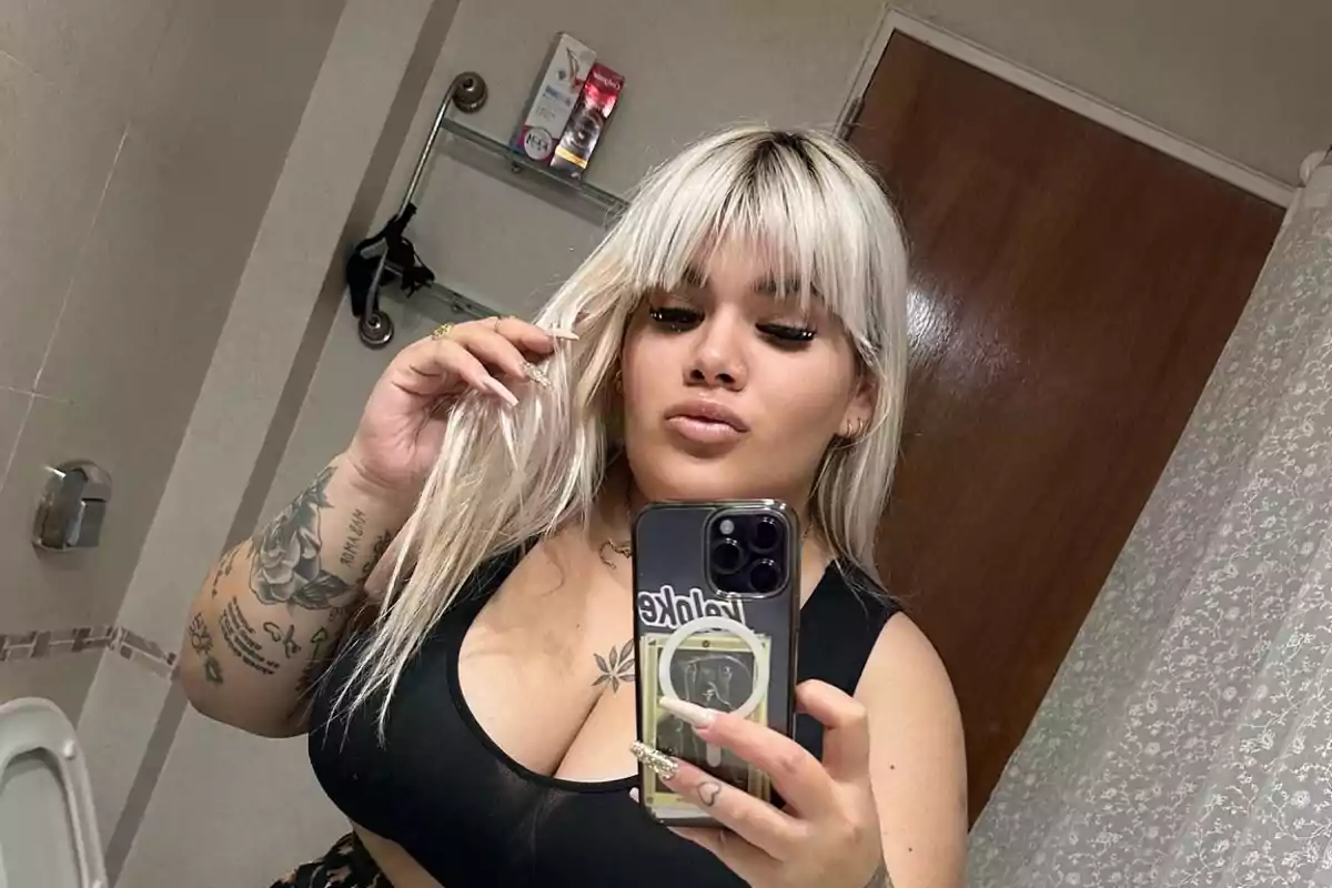 Persona con cabello rubio y tatuajes tomándose una selfie en un baño.