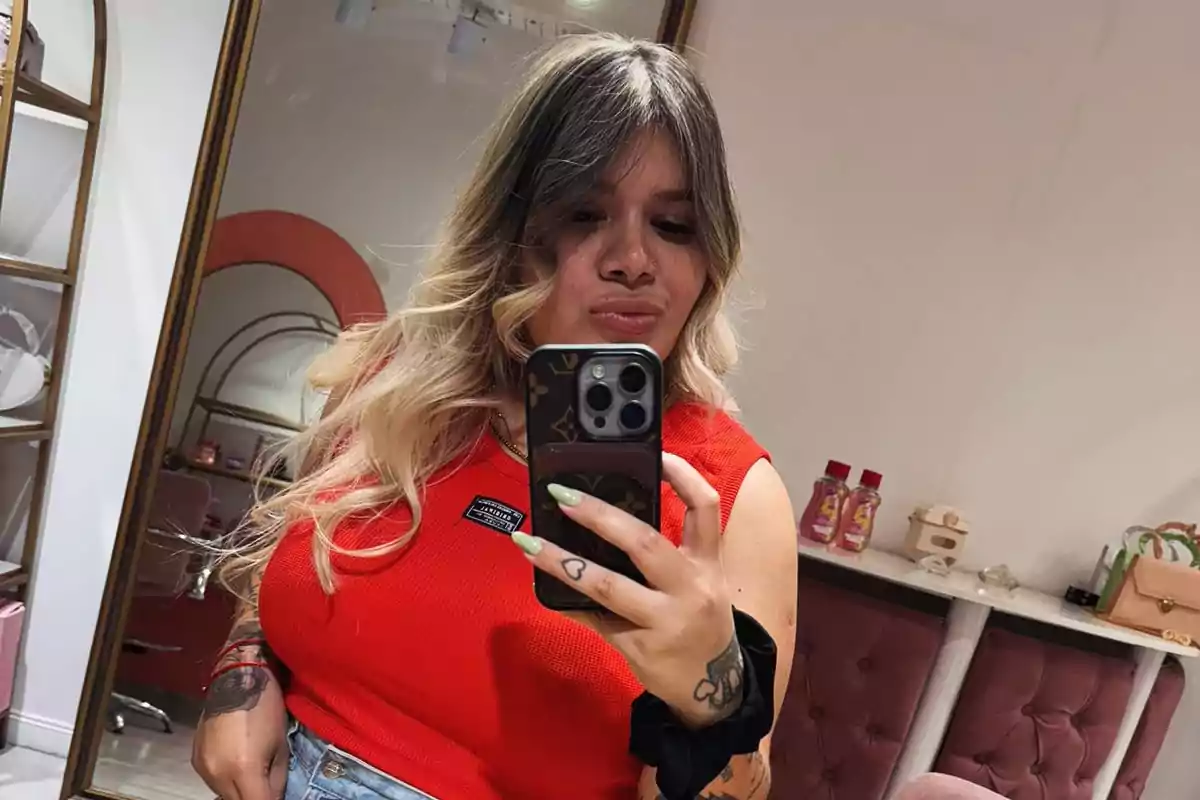 Una persona con cabello rubio y oscuro se toma una selfie frente a un espejo, vistiendo una camiseta roja y sosteniendo un teléfono móvil.