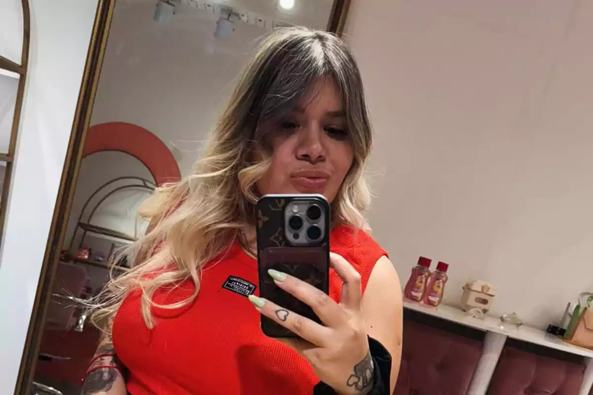 Una persona con cabello rubio y oscuro se toma una selfie frente a un espejo, vistiendo una camiseta roja y sosteniendo un teléfono móvil.