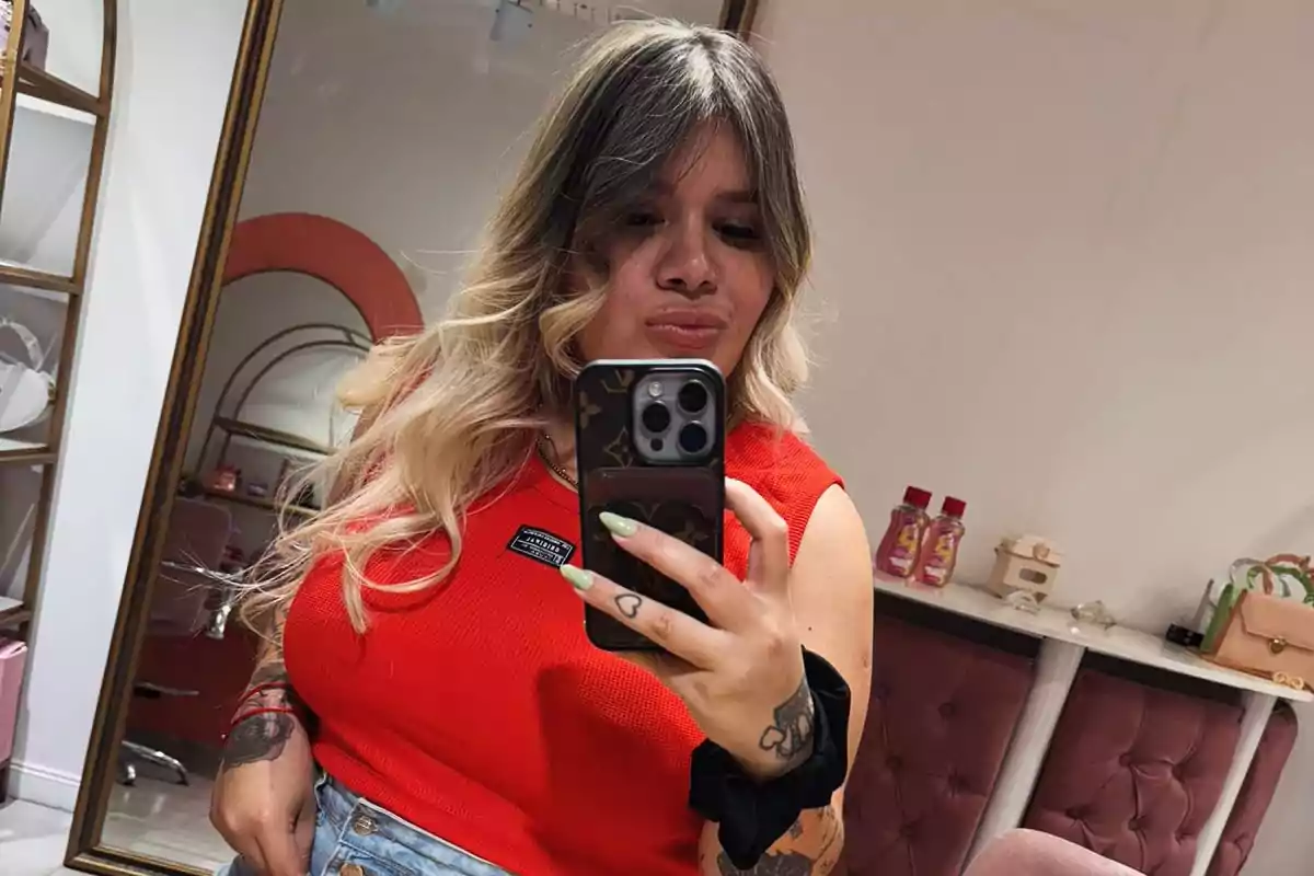 Una persona con cabello rubio y oscuro se toma una selfie frente a un espejo usando una camiseta roja y sosteniendo un teléfono móvil.