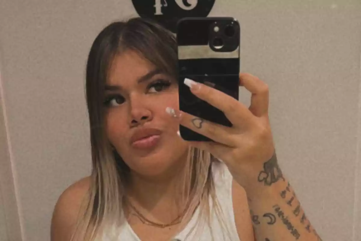 Una persona con cabello rubio y tatuajes en el brazo se toma una selfie frente a un espejo usando un teléfono móvil.