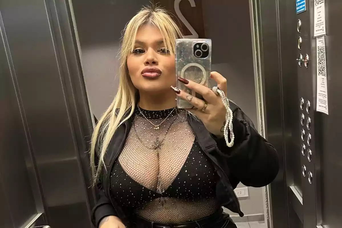 Persona rubia tomándose una selfie en un ascensor, usando una chaqueta negra y una blusa de malla con detalles brillantes. Persona rubia tomándose una selfie en un ascensor, usando una chaqueta negra y una blusa de malla con detalles brillantes.