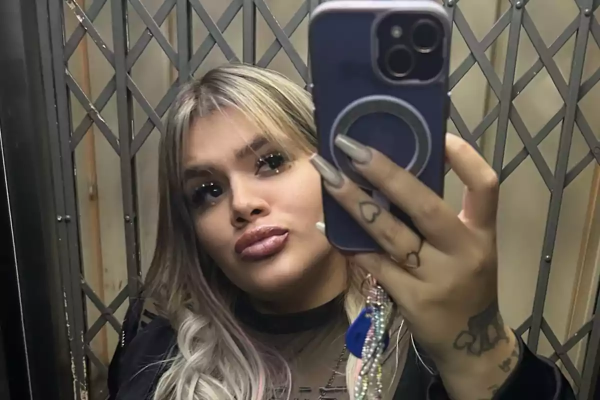 A person with blonde hair and long nails takes a selfie in a mirror using a mobile phone.