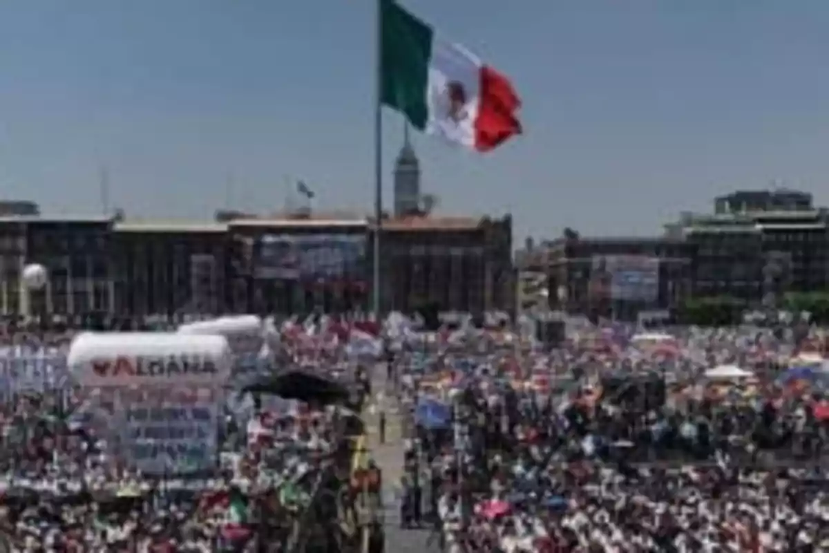 Una multitud se reúne en una plaza con una gran bandera de México ondeando en el centro.