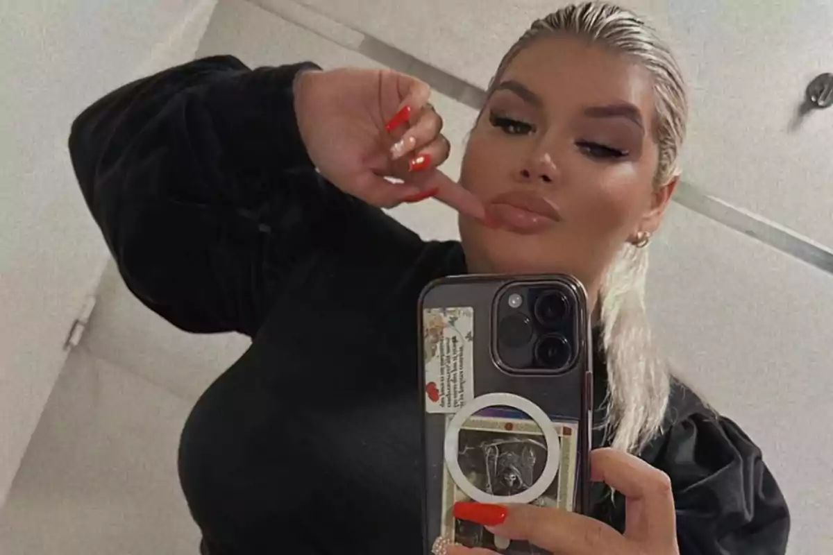 A person with blonde hair and red nails takes a selfie in front of a mirror using a mobile phone.