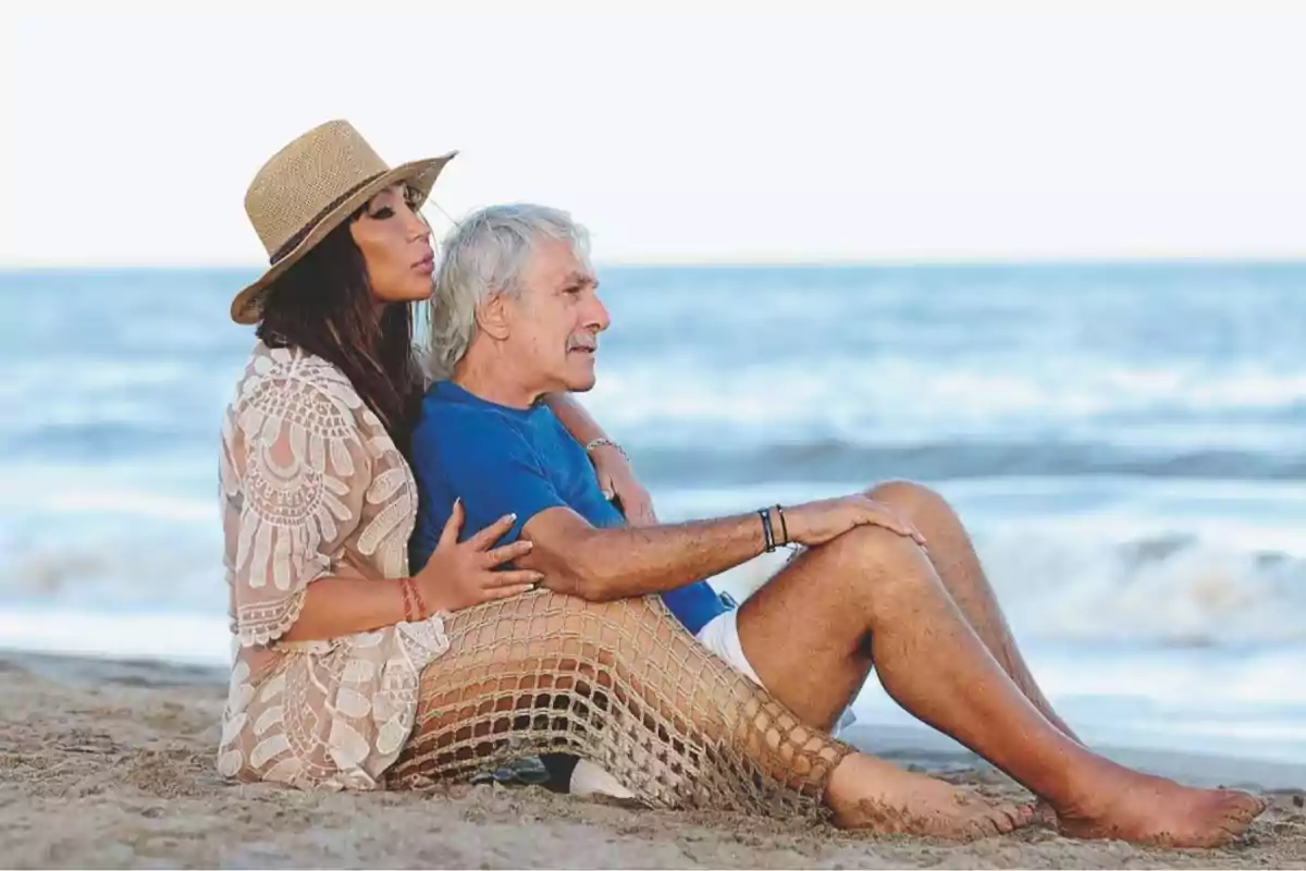 Una pareja sentada en la playa mirando el mar, la mujer lleva un sombrero y un vestido de encaje mientras el hombre viste una camiseta azul.