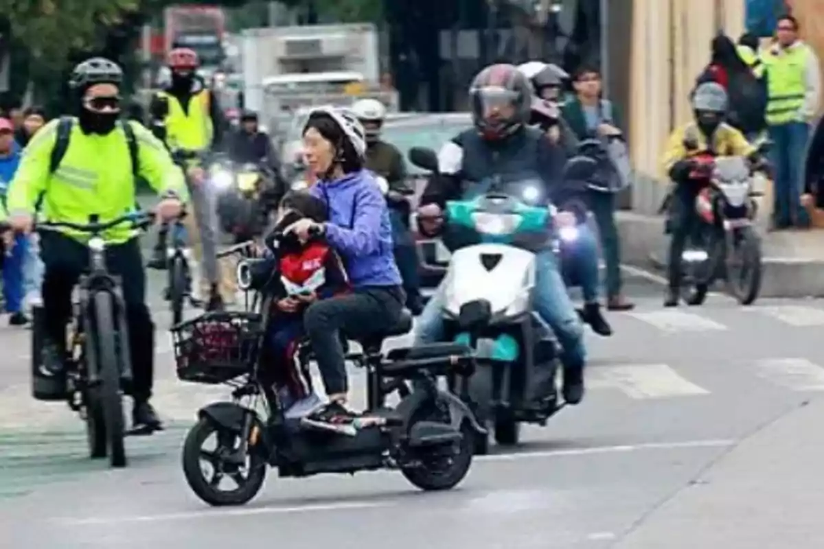 Una mujer y un niño viajan en un scooter eléctrico en una calle concurrida rodeados de ciclistas y motociclistas.