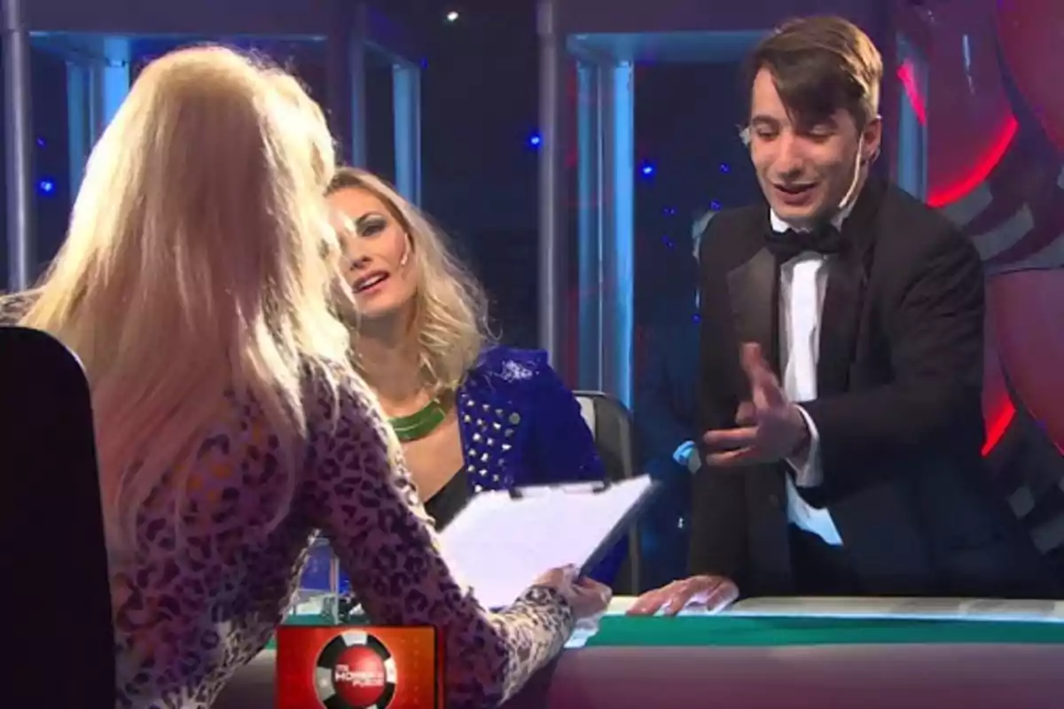 Tres personas están interactuando en un set de televisión, una de ellas sostiene un portapapeles mientras las otras dos parecen estar conversando animadamente. Tres personas están interactuando en un set de televisión, una de ellas sostiene un portapapeles mientras las otras dos parecen estar conversando animadamente.