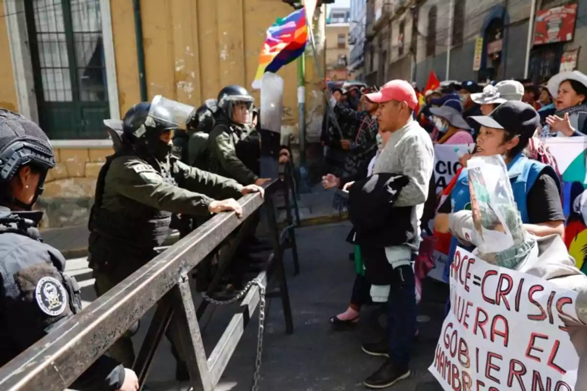 Policías con equipo antidisturbios forman una barrera frente a un grupo de manifestantes que sostienen pancartas y banderas multicolores en una calle urbana. Policías con equipo antidisturbios forman una barrera frente a un grupo de manifestantes que sostienen pancartas y banderas multicolores en una calle urbana.