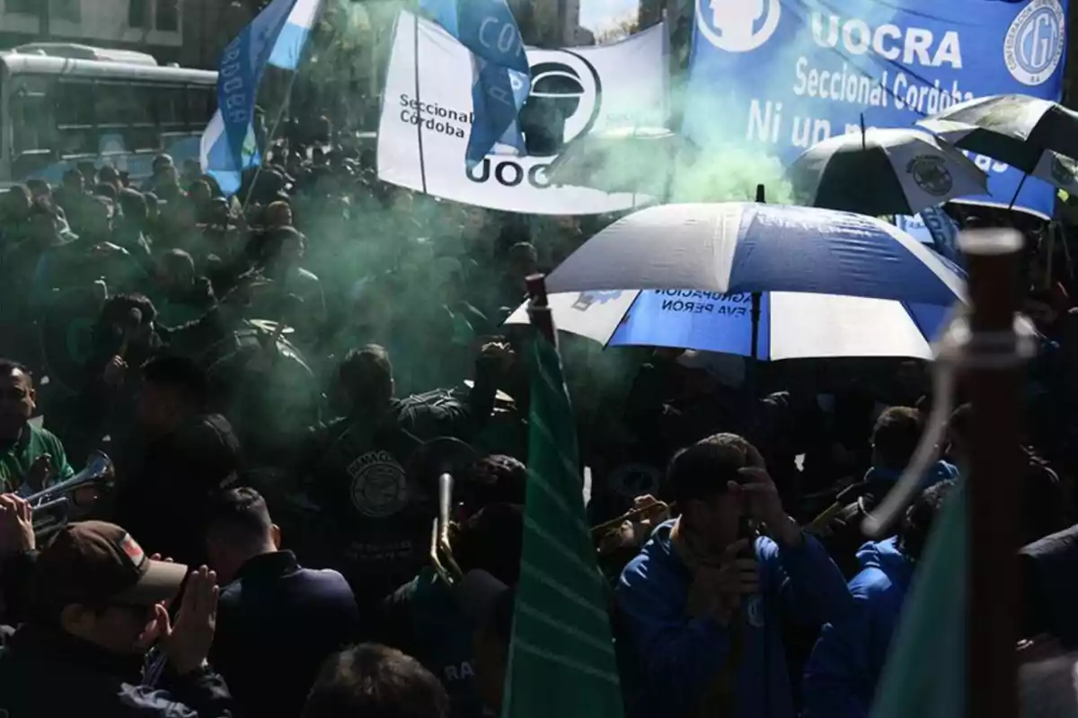 Manifestación de trabajadores con banderas y paraguas azules y verdes en una calle, acompañados de humo y pancartas de la UOCRA Seccional Córdoba Manifestación de trabajadores con banderas y paraguas azules y verdes en una calle, acompañados de humo y pancartas de la UOCRA Seccional Córdoba