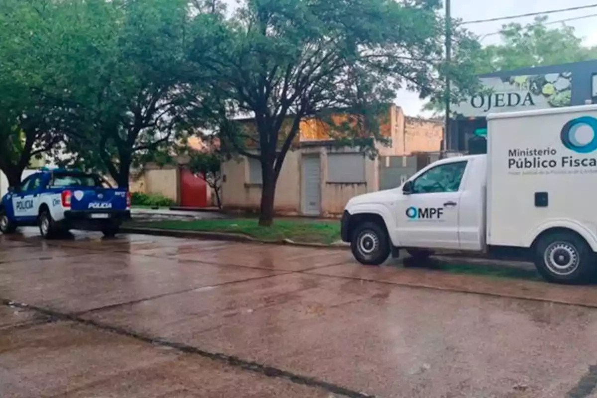 Dos camionetas oficiales, una de la policía y otra del Ministerio Público Fiscal, están estacionadas en una calle mojada frente a una casa y árboles. Dos camionetas oficiales, una de la policía y otra del Ministerio Público Fiscal, están estacionadas en una calle mojada frente a una casa y árboles.