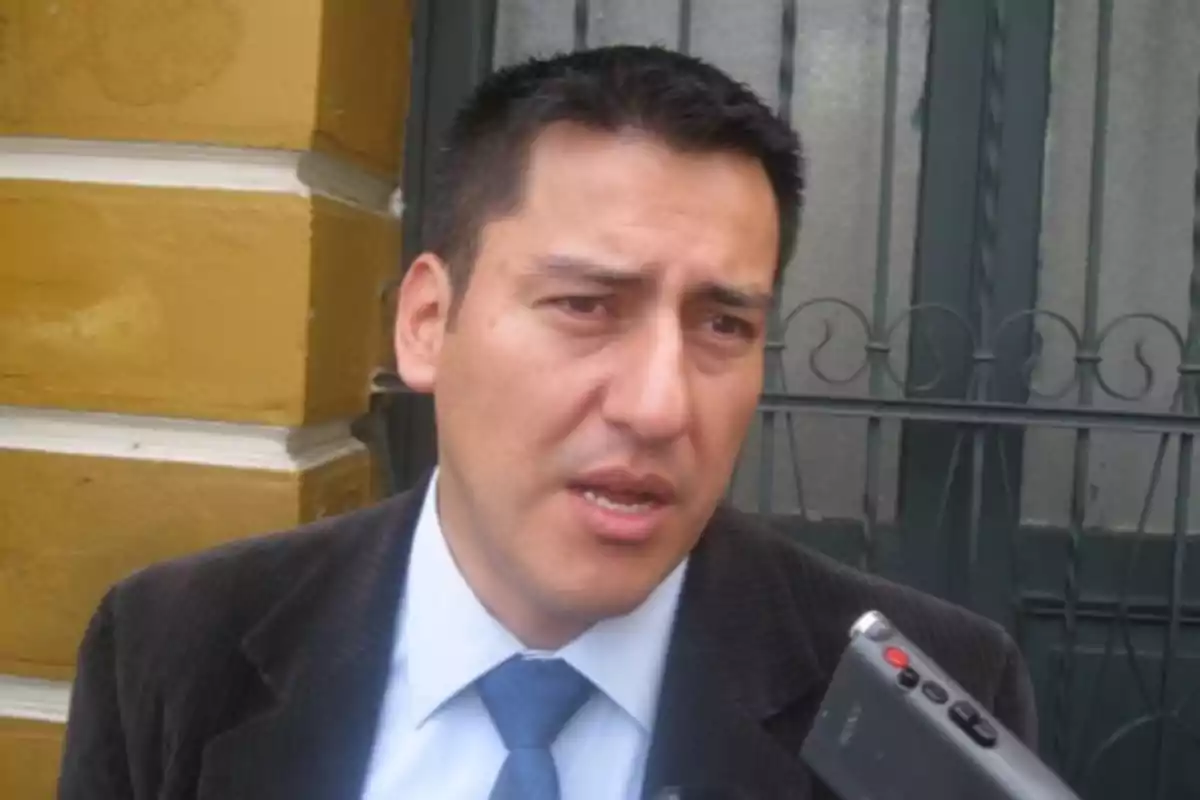 Hombre de cabello corto y traje oscuro hablando frente a un micrófono con una pared amarilla y una reja de fondo Hombre de cabello corto y traje oscuro hablando frente a un micrófono con una pared amarilla y una reja de fondo