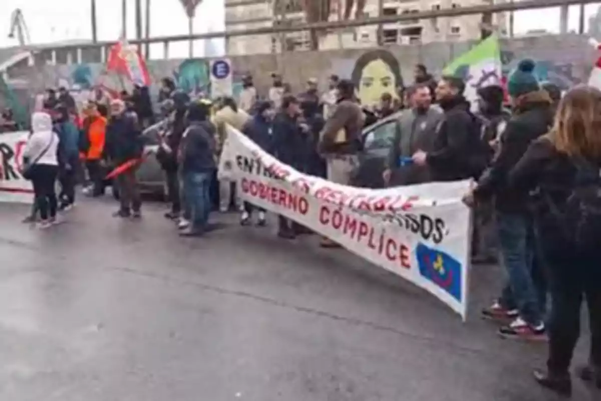 A group of people is protesting in the street holding signs and flags; one of the signs has red and black letters, and in the background, there is a mural with the face of a woman.