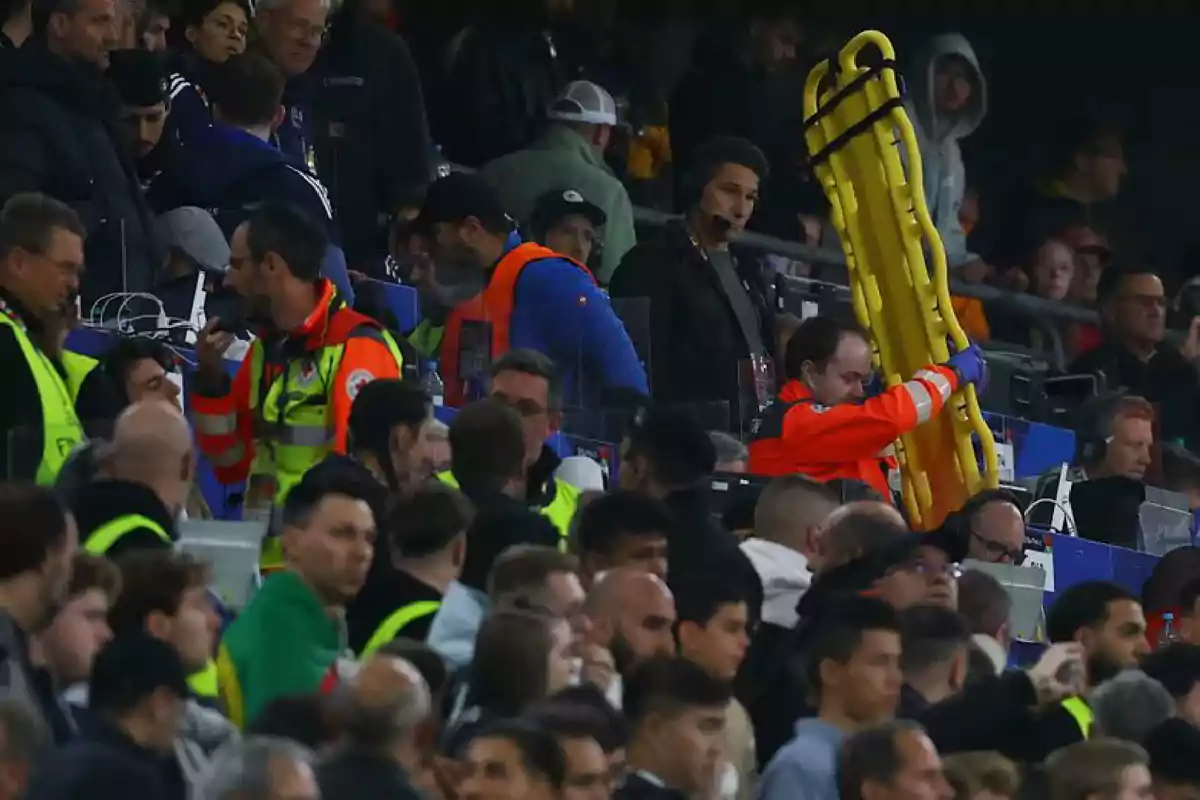 Personal de emergencia con uniforme naranja y chaleco amarillo se mueve entre la multitud en un estadio mientras uno de ellos lleva una camilla amarilla