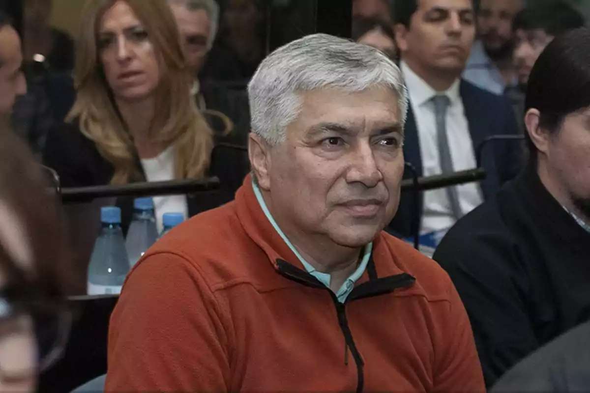 Hombre de cabello canoso con suéter naranja sentado entre otras personas en un ambiente formal