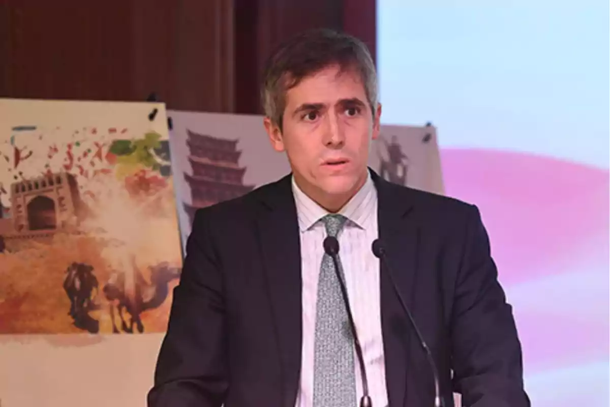 Hombre de traje hablando frente a dos micrófonos con ilustraciones coloridas al fondo Hombre de traje hablando frente a dos micrófonos con ilustraciones coloridas al fondo