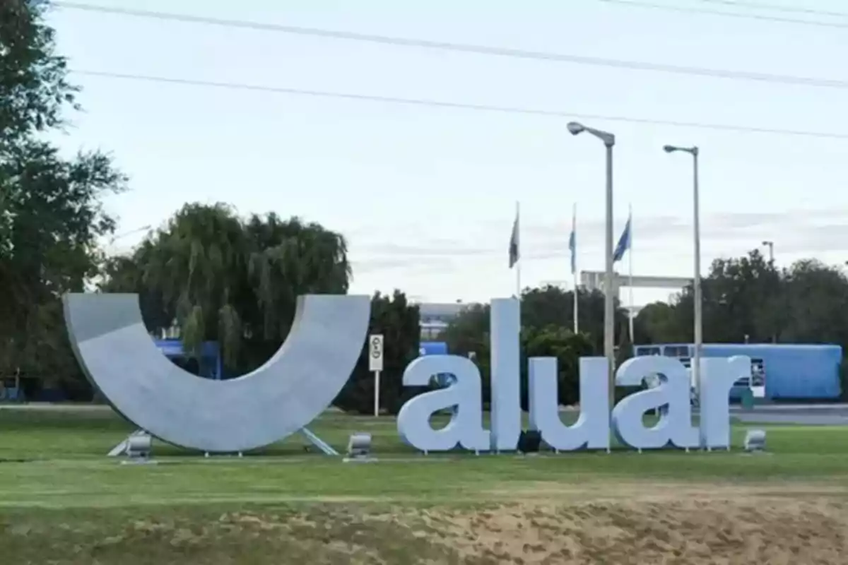 Un letrero grande de "aluar" en un área verde con árboles y banderas al fondo.