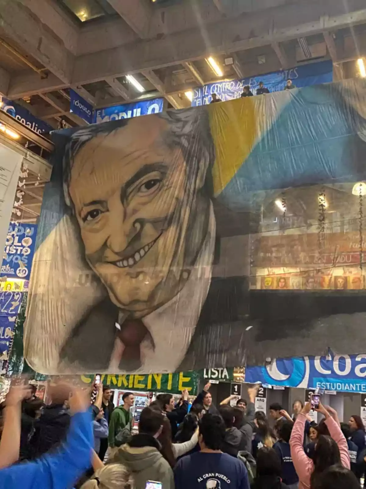 A group of people gathered in an enclosed space is holding a large flag with the portrait of a smiling man, while in the background there are posters and banners of different colors. A group of people gathered in an enclosed space is holding a large flag with the portrait of a smiling man, while in the background there are posters and banners of different colors.