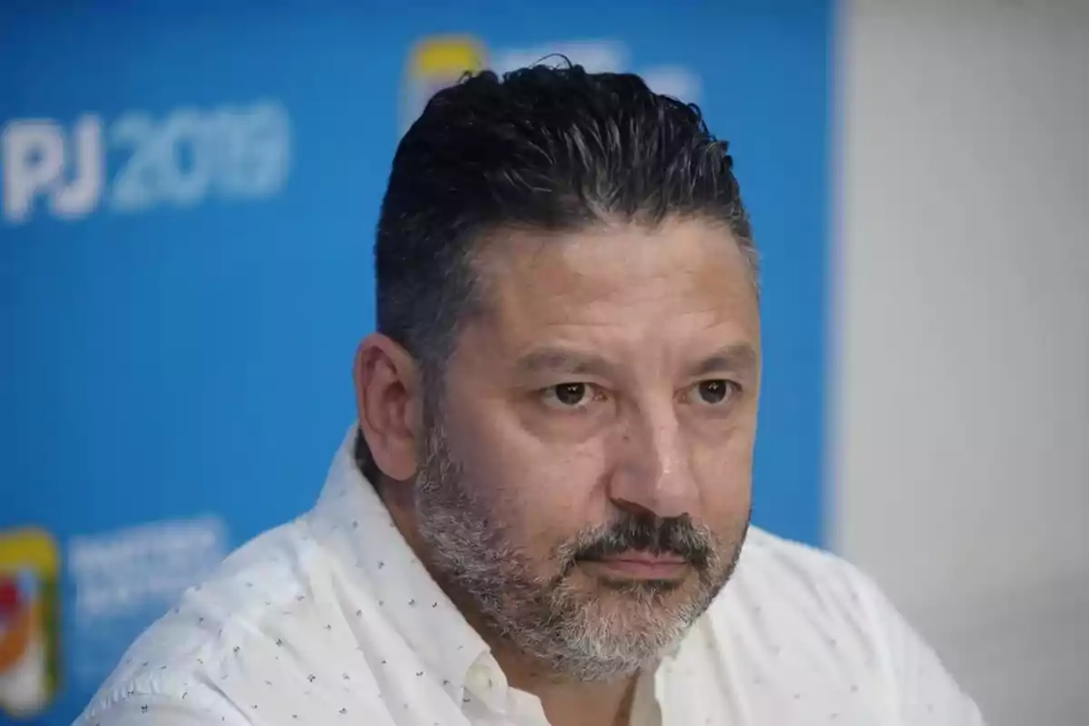 Un hombre con barba y cabello oscuro mira hacia adelante con expresión seria, frente a un fondo azul.